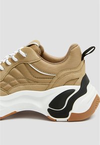 Baskets beige avec une tige en tissu texturé, des lacets blancs et une semelle épaisse blanche ornée d'accents noirs en caoutchouc.