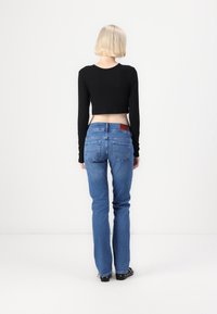 Personne aux cheveux blonds courts, portant un crop top noir à manches longues, un jean bleu et des chaussures noires, debout de dos sur un fond uni.