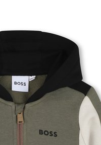 Kurtka z kapturem i zamkiem, charakteryzująca się czarnym kapturem, oliwkowym krojem i białymi rękawami. Posiada prążkowaną teksturę oraz wyraźne logo 'BOSS'.