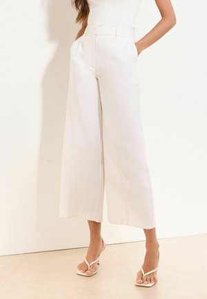 Vrouw die witte, hooggetailleerde, wijd uitlopende broek en witte sandaaltjes met hak en bandjes draagt, staand met handen in de zakken.