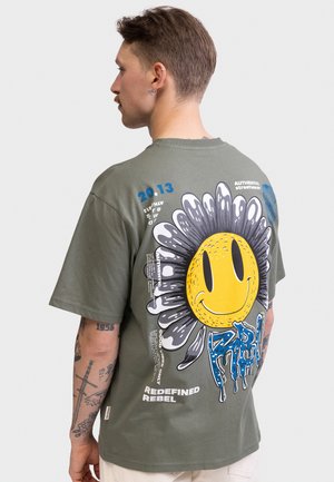 Mand med øre-piercinger og tatoveringer iført en olivengrøn t-shirt med en stor smilende blomst og teksten "REDEFINED REBEL" på ryggen.
