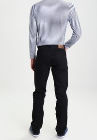 Persona con jeans negros, camisa de rayas blanca y azul marino de manga larga, y zapatillas blancas, de pie con una mano detrás de la espalda.