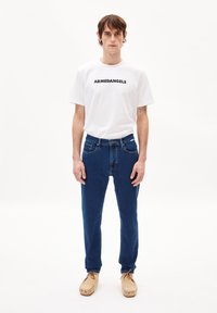 Camiseta blanca de manga corta con la marca "ARMEDANGELS" en negro, combinada con jeans de mezclilla azules y zapatos estilo mocasín en color beige.