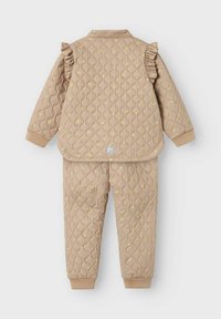 Quiltet beige jakke med volanger ved skuldrene og blomsterprint, parret med matchende bukser. Accenter inkluderer elastiske manchetter og subtile teksturer.