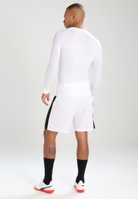 Haut de sport à manches longues blanc, ajusté ; associé à un short blanc avec des rayures noires sur les côtés. Les accessoires incluent des chaussettes noires jusqu'aux genoux et des chaussures.