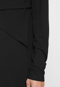 Sort, ribstrikket langærmet beklædningsgenstand med et lagdelt, asymmetrisk detalje over taljen og en hånd synlig ved manchet.