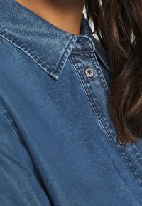 Jeansskjorta i mörkblå färg, med spetsig krage, knappstängning och texturerad yta. Synlig knappdetalj vid halsen.