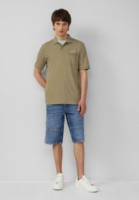 Camisa de polo verde oliva de manga corta con un bolsillo en el pecho y diseño bordado, combinada con pantalones cortos de mezclilla y zapatillas blancas.
