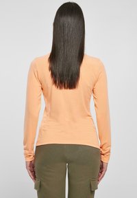 Maglietta arancione a maniche lunghe realizzata in tessuto morbido, caratterizzata da un design aderente e un orlo arrotondato. Abbinata a pantaloni cargo verde oliva.