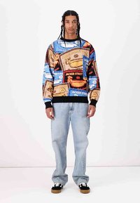 Multicolored sweatshirt with graphic patterns featuring "Jacker Street" and various designs. Black cuffs and neckline, worn with light jeans and sneakers.