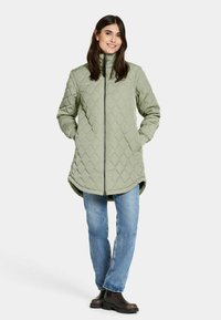 Lichtgroene gewatteerde jas met hoge kraag en rits, voorzien van een ruitpatroon en twee zijzakken, gecombineerd met blauwe jeans en bruine laarzen.