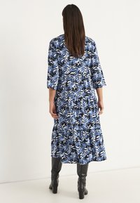 Blumenkleid Maxi in Blau und Schwarz mit 3/4-Ärmeln, gestuften Design und fließender Silhouette. Kombiniert mit knielangen schwarzen Stiefeln.