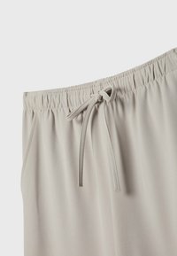 Pantalon beige clair à taille élastique avec un cordon noué et des poches latérales visibles sur un fond uni.