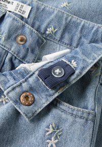 Svetlo modra denim tkanina z vezenimi belimi cvetovi. Vsebuje rjav kovinski gumb in mornarsko modre tkanine poudarek s prozornim gumbom. Vidna je etiketa za nego.