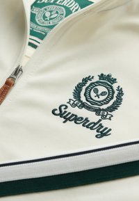 Witte jas met ritssluiting, voorzien van een donker groene geborduurde logo en accenten, met een gestructureerde stof en een gestreept detail bij de zoom.