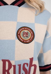 Nærbillede af blå og cremefarvet ternet sweater med et rundt patch, der siger "Forbudt Motorsport Company Berlin Tyskland" og røde tekstdele, der er delvist synlige.