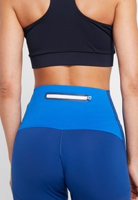 Gros plan sur le dos d’une personne portant un soutien-gorge de sport noir et des leggings bleus taille haute avec une petite poche zippée à la taille.