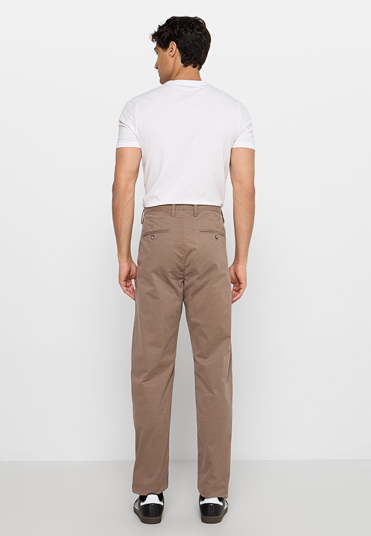 Braune Baumwollhose, entspannte Passform, mit Gesäßtaschen und sichtbaren Steppnähten. Kombiniert mit einem schlichten weißen T-Shirt und schwarzen Turnschuhen.