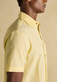 Mężczyzna w krótkim, jasnożółtym koszuli na guziki, pokazany z boku na neutralnym tle.