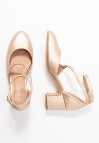 Glänzende beige Damenblockabsatzschuhe mit Knöchelriemen, ein Schuh steht aufrecht, der andere liegt auf der Seite, vor einem weißen Hintergrund.