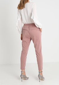 Pantalons ajustés rose clair avec une jambe effilée, une taille élastique et deux poches arrière, associés avec un blouse à manches longues blanches à volants et des talons argentés.