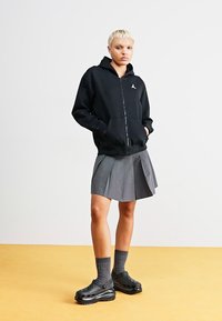 Sudadera negra con cremallera y logo, falda gris con plisado, calcetas grises hasta la rodilla y zuecos negros sobre un suelo amarillo con un fondo liso.