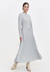 Frau trägt ein langärmliges, hellgraues, plissiertes Midikleid, kombiniert mit cremefarbenen Stiefeletten und blumigen Ohrringen, lächelnd stehend vor einem weißen Hintergrund.