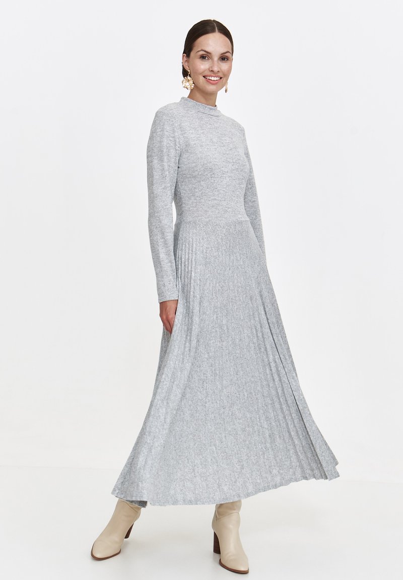 Frau trägt ein langärmliges, hellgraues, plissiertes Midikleid, kombiniert mit cremefarbenen Stiefeletten und blumigen Ohrringen, lächelnd stehend vor einem weißen Hintergrund.