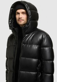 Giacca a vento nera in materiale lucido con un grande cappuccio strutturato. Presenta una chiusura con zip e tasche laterali, evidenziando un design trapuntato.