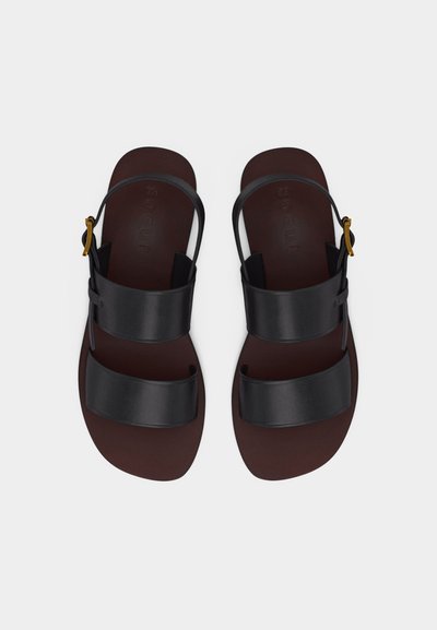 Ζευγάρι σκούρα καφέ φλατ σανδάλια με δύο πλαίες μαύρες δερμάτινες λωρίδες και λουράκι αστραγάλου με χρυσές αγκράφες.