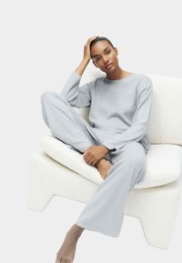 Grijze geribbelde loungewear set met lange mouwen en wijde broekspijpen, zittend op een textuur witte stoel. Soepele textuur, ontspannen pasvorm.