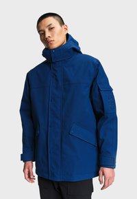 Homme portant une veste à capuche bleu foncé avec des fermetures à boutons-pression et des poches à rabat, debout devant un fond clair uni.