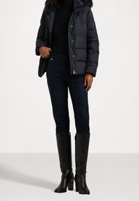 Padded tmavě modrá bunda s kapucí, černý rolák, obtažené tmavé kalhoty a černé kozačky na podpatku do kolen. Měkká textura, detaily se zipem.