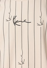 T-Shirt beige con strisce nere verticali, caratterizzato da disegni a linee astratte e una firma al centro. Tessuto morbido con texture liscia.