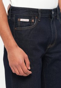Tamne traperice tamne boje s narančastim šavovima, prednji džep s bijelom i crvenom oznakom Calvin Klein, u klasičnom ravnom kroju.