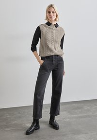 Ljusbeige kabelstickad ärmlös tröja över en svart långärmad skjorta, i kombination med högmidjade svarta jeans och ankellångstövlar.