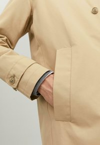 Lichtbeige jas van gladde stof, met een met knopen verstelbare manchette en een zijzak. De zichtbare binnenlaag is een grijze gebreide trui.