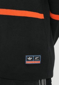 Črn pleteni pulover z oranžno vodoravno črto. Na spodnjem levem delu se nahaja logo nalepka z blagovnima znamkama Adidas in All Blacks.