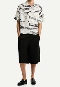 Lyhythihainen paita, jossa on mustavalkoinen abstrakti kuvio, yhdistetty polvipituisen mustien shortsien ja mustien loaferien kanssa.