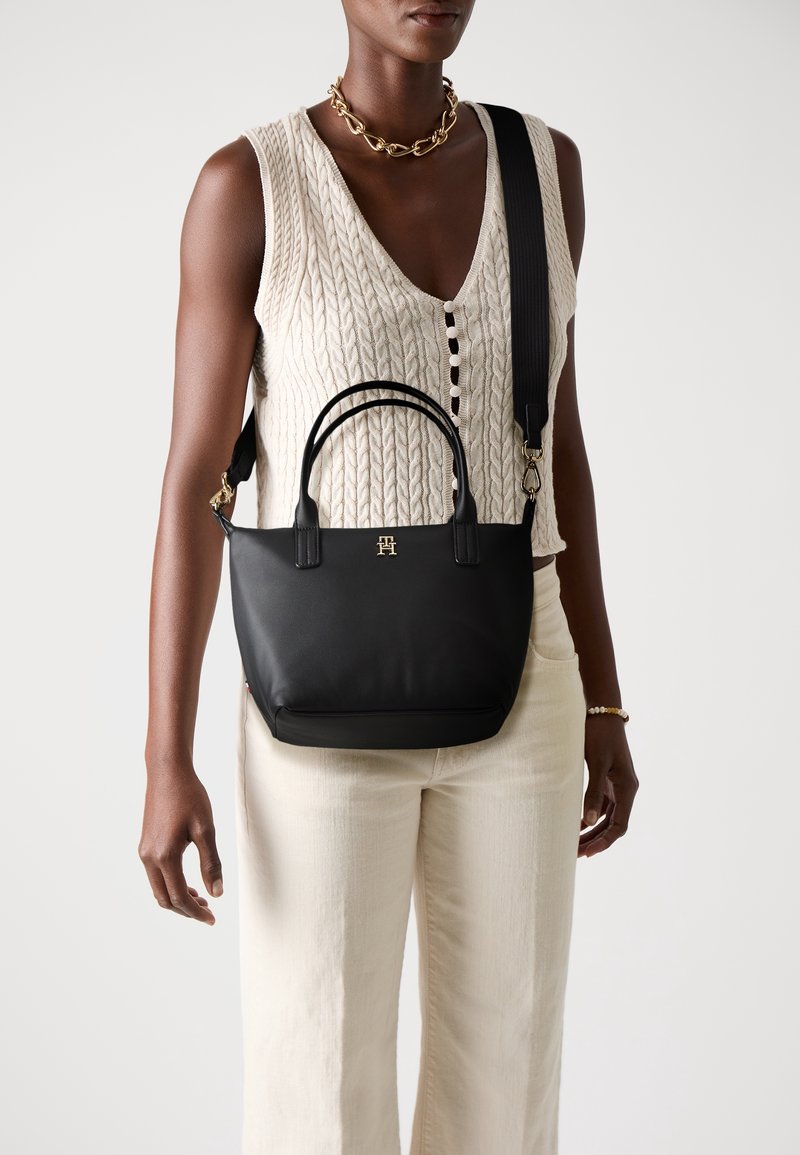 Sac à main en cuir noir avec doubles poignées et large bandoulière porté en travers par une personne portant un gilet torsadé crème et un pantalon.