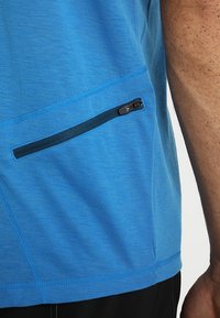 Modrá sportovní tričko s hladkou texturou, obsahující zipovou kapsu na boku a kontrastní prošívání.