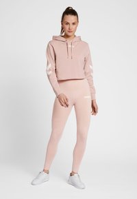 Różowa krótka bluza z kapturem z białym logo i akcentami w kształcie szewronu, zestawiona z dopasowanymi legginsami z wysokim stanem ozdobionymi logo oraz białe trampki.