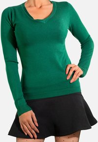 Pull en maille verte avec un décolleté en V et des manches longues, fabriqué à partir de matériau doux, associé à une jupe noire évasée.