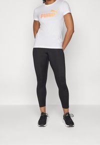 Camiseta blanca con el logo naranja de Puma, combinada con leggings negros. Zapatillas de deporte negras con acentos amarillos. Ajuste moderno, diseño minimalista.