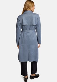Manteau bleu clair en suédine synthétique avec une taille ceinturée, un détail à rabat dans le dos, des manches longues et une coupe mi-longue, porté avec un pantalon foncé.
