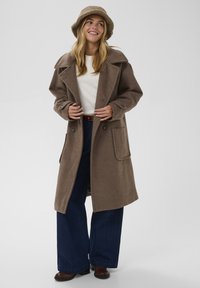 Rjav plašč iz volnene mešanice z velikimi klapnicami, gumbastim zapiranjem in dvema sprednjima žepoma. V kombinaciji z kremno majico in širokima temno modrima hlačami.