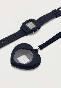 Ψηφιακό ρολόι χειρός Casio σε ναυτικό μπλε δίπλα σε μεγεθυντικό φακό σχήματος καρδιάς σε ναυτικό μπλε με λουράκι σε λευκό φόντο.