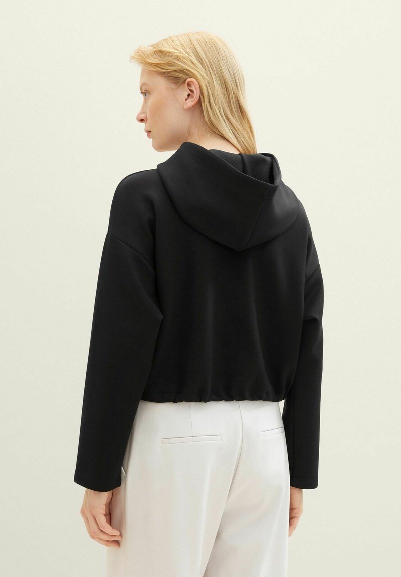 Schwarzer, cropped Hoodie mit strukturiertem Finish, verstellbarer Kapuze und langen Ärmeln, kombiniert mit hellen Hosen.