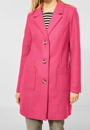Femme portant un manteau rose vif à simple boutonnage avec de grandes poches à l'avant, porté sur une chemise rayée noire et blanche et un pantalon gris.