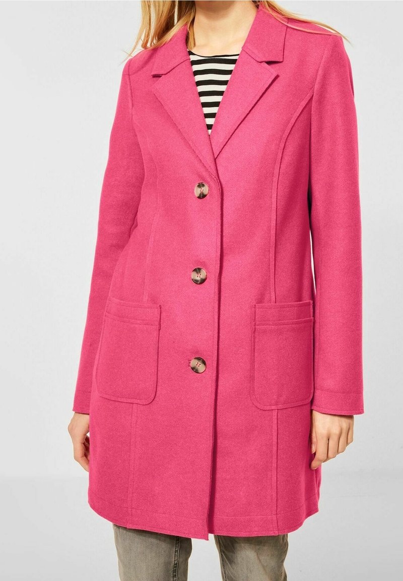Femme portant un manteau rose vif à simple boutonnage avec de grandes poches à l'avant, porté sur une chemise rayée noire et blanche et un pantalon gris.